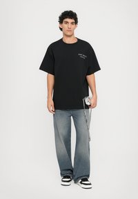 Svart oversized t-skjorte med korte ermer som har hvit tekst, sammen med lyseblå baggy jeans og svart-hvite sneakers. Sølvkjede detalj.
