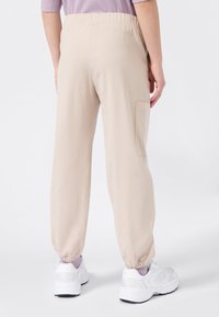 Personne portant un pantalon de jogging beige avec des chevilles élastiques, une poche latérale, des baskets blanches et un haut lavande, se tenant devant un fond blanc.