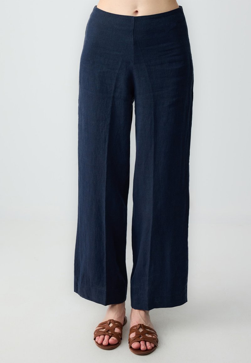 Marineblauwe linnen wijde broek met hoge taille en relaxte pasvorm. Glad textuur, geen zichtbare patronen. G gedragen met bruine sandaaltjes met bandjes.