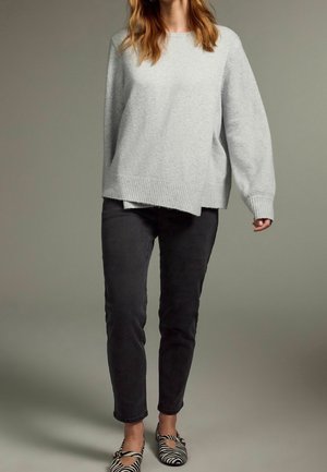 Femme portant un pull oversize gris clair, un jean noir slim et des ballerines à imprimé zèbre noir et blanc, debout sur un fond gris uni.