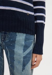 Μπλε ναυτικό ριγέ πουλόβερ με ριμπ τελείωμα και μανσέτες, με ανοιχτό μπλε και λευκές οριζόντιες ρίγες. Φορεμένο πάνω από σκισμένο μπλε τζιν.