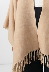 Beige strikket poncho med frynset kant, som har en myk tekstur og åpen frontdesign. Egnet for lag-på-lag over uformelle antrekk.