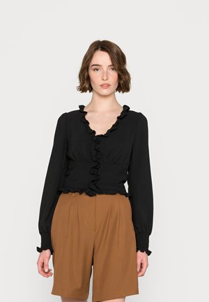 Blusa preta com volantes, mangas longas, decote em V e cintura franzida, combinada com calções pregueados castanhos. Textura do tecido suave.