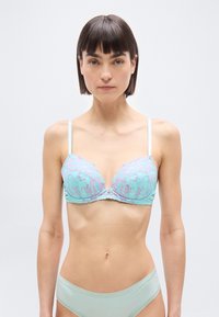 Γυναίκα που φοράει ανοιχτό μπλε δαντελωτό σουτιέν με ροζ λουλουδένιες λεπτομέρειες και ταιριαστό εσώρουχο, στέκεται μπροστά σε απλό λευκό φόντο.