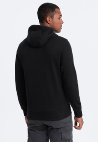 Sudadera negra con capucha ajustable, puños y dobladillo acanalados. Textura suave, ajuste cómodo y diseño minimalista, vista desde la parte de atrás.