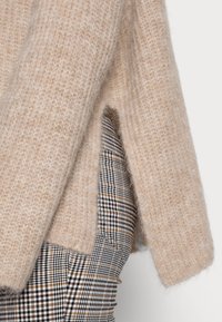 Maglione lavorato a maglia beige con una finitura morbida e testurizzata e apertura laterale, sovrapposto a un tessuto a quadri bianco e nero.
