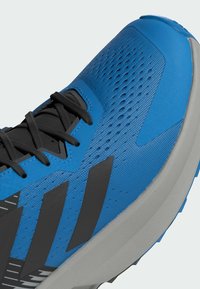 Blå sportskor med en texturerad yta, svarta accenter och perforeringar. Har en dämpad sula och svarta snören.