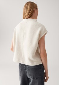 Vrouw met schouderlang blond haar, gekleed in een mouwloze witte gebreide top met textuur en donkere grijze jeans, van achteraf zichtbaar.