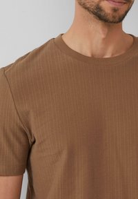 Braunes, geripptes T-Shirt mit Rundhalsausschnitt, kurzärmlig und aus strukturierter Stoffqualität. Nahaufnahme, die die Nähte und den Ausschnitt zeigt.