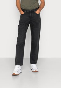 Jeans de mezclilla negro con un corte recto, complementados con una camiseta verde y zapatillas blancas con suelas de color marrón claro.