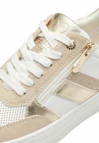 Bež ja valge sneaker, millel on metallik kuldsed aktsendid, perforeeritud detail, valged paelad ja külgipaigaldus, millel on südamekujuline tõmblukk.