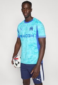 Φωτεινό γαλάζιο ποδοσφαιρικό φανελάκι με κοντά μανίκια και μπλε λεπτομέρειες, που φέρει σχέδιο με αξιοθέατα και λογότυπα. Μαύρη και λευκή ποδοσφαιρική μπάλα κρατημένη στο ένα χέρι.