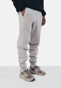 Pantalons de survêtement gris avec des poignets élastiques, poches avant et logo orange brodé. Associés à des chaussures de sport beige et grises.