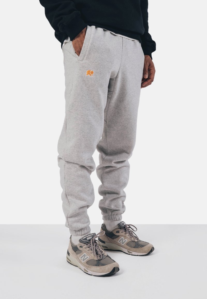 Pantalons de survêtement gris avec des poignets élastiques, poches avant et logo orange brodé. Associés à des chaussures de sport beige et grises.