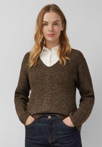 Kobieta z blond włosami ubrana w brązowy sweter w serek na białej koszuli z kołnierzykiem oraz ciemnoniebieskich dżinsach, z rękami w kieszeniach.