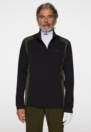 Mand iført sort og olivengrøn quarter-zip trøje, hvid turtleneck med sort logo, olivengrønne bukser og en hvid handske på højre hånd.