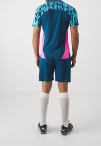 Sportovní dres s modro-turquoise geometrickým designem, růžovými bočními akcenty, spárovaný s modrými šortkami a bílými ponožkami až pod kolena.