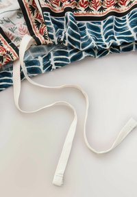 Tissu avec bordure florale rouge, noire et crème et motif abstrait bleu, présentant de longs cordons blanc cassé sur un fond neutre.