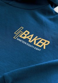 Blauwe hoodie met "BAKER" in vetgouden letters en "NINETEEN EIGHTY EIGHT" in het wit, met een gestructureerde stofafwerking.