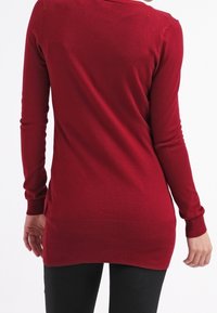 Roter Strickpullover mit langen Ärmeln, rundem Ausschnitt und geripptem Saum. Besitzt eine glatte Textur und eine figurnahe Passform, getragen über schwarzen Leggings.