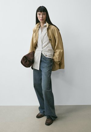 Frau steht vor schlichter Wand, trägt beige Jacke, weißes gestreiftes Hemd, lockere blaue Jeans und braune Schuhe, hält gefalteten braunen Hut in der Hand.