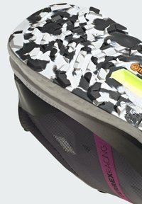 Zapatilla de trail running con una suela texturizada en blanco y negro, acentos morados y parte superior de malla transpirable, diseñada para ofrecer agarre y soporte.