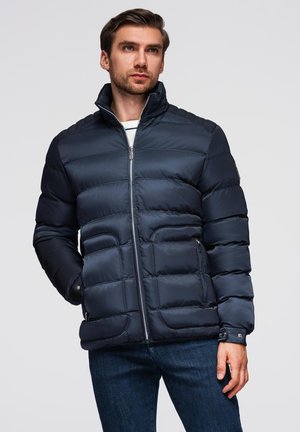 Giacca puffer blu navy con colletto alto, chiusura con zip, trapuntatura orizzontale e due tasche laterali. Realizzata in un tessuto liscio e isolante.