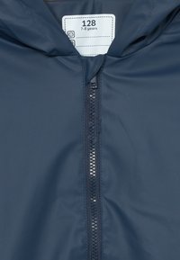 Dunkelblaue Jacke mit einem Reißverschluss vorne, Kapuze und einem Etikett, das die Größe 128 für 7-8 Jahre angibt. Glatte Textur und leichtes Material.