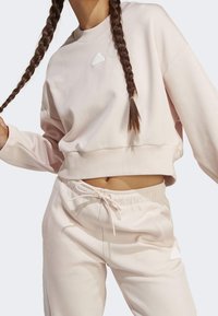 Ljusrosa sweatshirt med rund halsringning och vit Adidas-logotyp. Matchas med joggerbyxor i samma färg med dragsko i midjan och sidofickor.