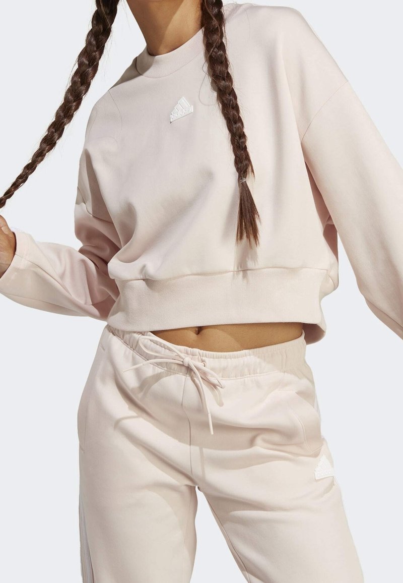 Ljusrosa sweatshirt med rund halsringning och vit Adidas-logotyp. Matchas med joggerbyxor i samma färg med dragsko i midjan och sidofickor.