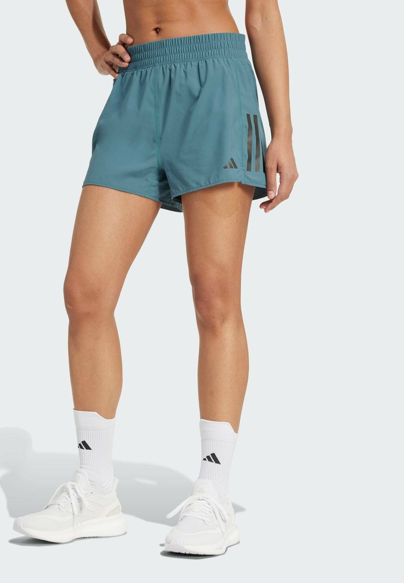 Türkise Sportshorts mit einem dehnbaren Gummi, die an der Seite drei schwarze Streifen haben. Kombiniert mit weißen Turnschuhen und Socken.