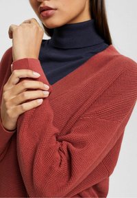 Maglione rosso strutturato con scollatura a V sovrapposto a un dolcevita blu scuro. Primo piano delle maniche e delle mani che tengono il tessuto.
