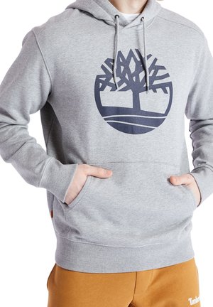 Osoba nosząca jasnoszarą bluzę z kapturem z dużym ciemnoniebieskim, stylizowanym logo drzewa na piersi, ręce w przedniej kieszeni oraz spodnie w kolorze musztardowym.