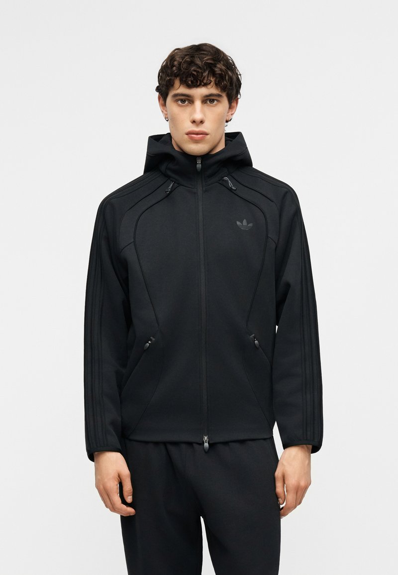 Jonge man met kort krullend haar, gekleed in een zwarte hoodie met rits en bijpassende zwarte broek, die recht vooruit kijkt tegen een effen achtergrond.