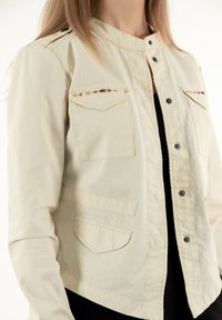 Giacca beige chiaro con chiusura a bottoni, due tasche sul petto con cuciture decorative e tasche laterali inclinate. Texture liscia.