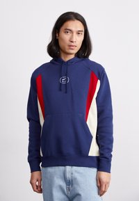 Hoodie bleu marine avec une poche frontale, présentant des panneaux latéraux rouges et crème ainsi qu'un logo sur la poitrine. Confectionné en tissu en mélange de coton.