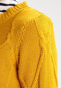 Stickad tröja i klar gult med en strukturerad mönster och ribbad rund halsringning. Har kabelstickad detalj längs axlarna.