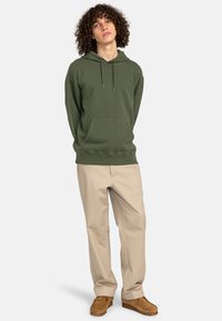 Olivegrøn hoodie med en frontlomme, snørret hætte og ribbet kant. Sammen med beige bukser og brune ruskindssko.