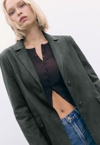 Mørkegrøn suedeblazer med en klapkrave, knaplukning foran, båret over en ribbet brun top, parret med højtsiddende blå jeans.