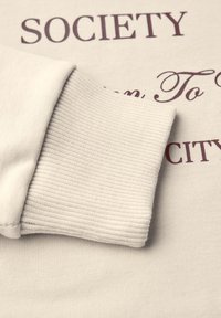 Felpa beige chiaro con polsini a coste e stampa di testo in maroon scuro. La texture del tessuto è liscia con una finitura morbida.