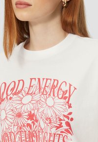 Witte katoenen t-shirt met een roze bloemen grafisch ontwerp en de tekst "GOEDE ENERGIE" en "GOEDE GEDICHTEN" in vetgedrukte typografie.