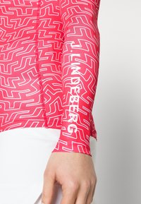 Närbild av en hand som bär en röd ärm med vita abstrakta linjer och texten "J. LINDEBERG" i vitt längs armen.