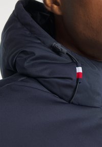 Marineblå hettegenser med glatt, strukturert stoff, justerbar snor og en liten trikolor logoakcent nær nakken.