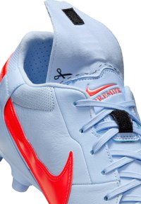 Világoskék focicipő piros Nike logóval, textúrázott bőrből készült és fekete tépőzárral. Kiemelkedő fűzőrendszerrel és perforált részekkel rendelkezik.