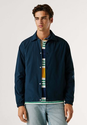 Chaqueta ligera azul marino con cierre de botones y puños elásticos, usada sobre una camisa de rayas multicolor. Estilo casual, textura suave.