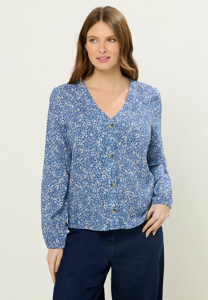 Femme aux cheveux châtain clair portant un chemisier bleu à motifs floraux boutonné et un pantalon bleu foncé, debout avec une main sur la hanche.
