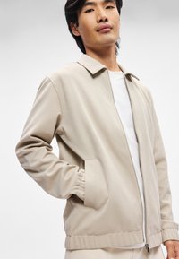 Beżowa kurtka zapinana na zamek z płaskim kołnierzem, elastycznymi mankietami i bocznymi kieszeniami, wykonana z miękkiego, gładkiego materiału, zaprezentowana na białej koszuli.