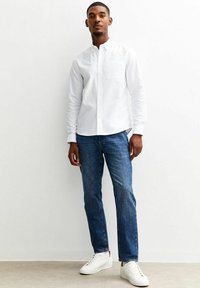 Camicetta bianca a maniche lunghe con colletto a bottoni, taschino singolo, abbinata a jeans blu e sneakers bianche su uno sfondo neutro.