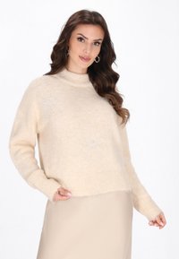 Pulover bej, pufos, cu guler înalt și maneci lungi. Prezintă motive florale din cristale pe față și manșete cu nervuri.