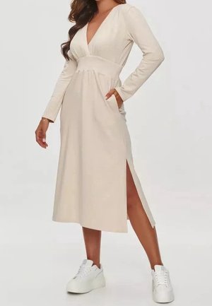 Mujer con vestido midi beige de manga larga con abertura lateral y zapatillas blancas, de pie con una mano en el bolsillo y la otra relajada.